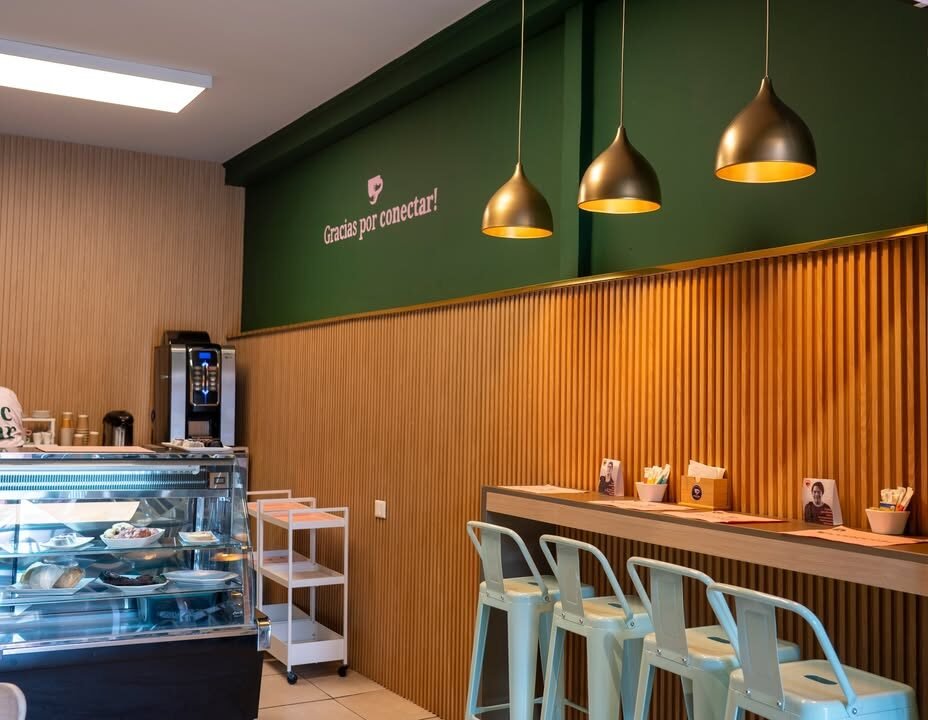 Interior of a café with a wood-slat counter and wall, a green upper wall reading 'Gracias por conectar!' in white script, and three gold dome pendant lights hanging above a bar-height counter lined with light blue metal stools. On the left, a glass display case holds pastries beside a coffee machine and a white utility cart.