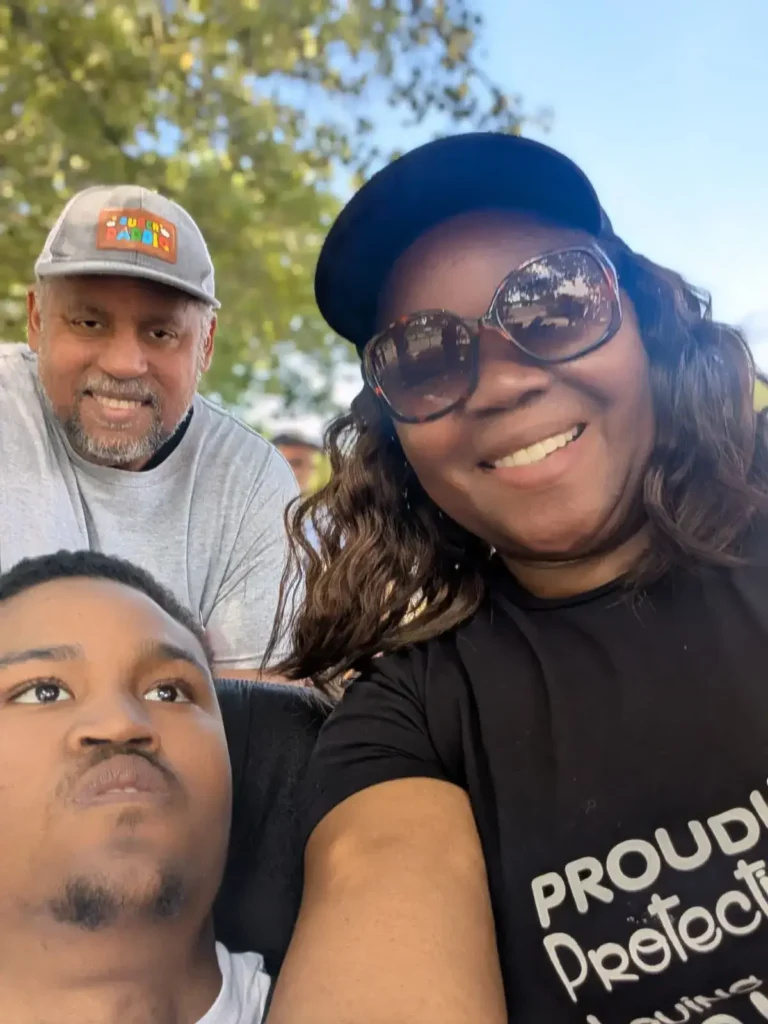 A selfie of a smiling woman in sunglasses, a young man, and an older man wearing a baseball cap at an outdoor event.