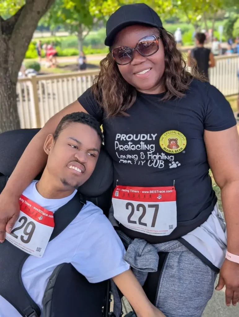 A smiling woman wearing sunglasses and a race bib stands with her arm around a young man in a wheelchair wearing a matching race bib outdoors.