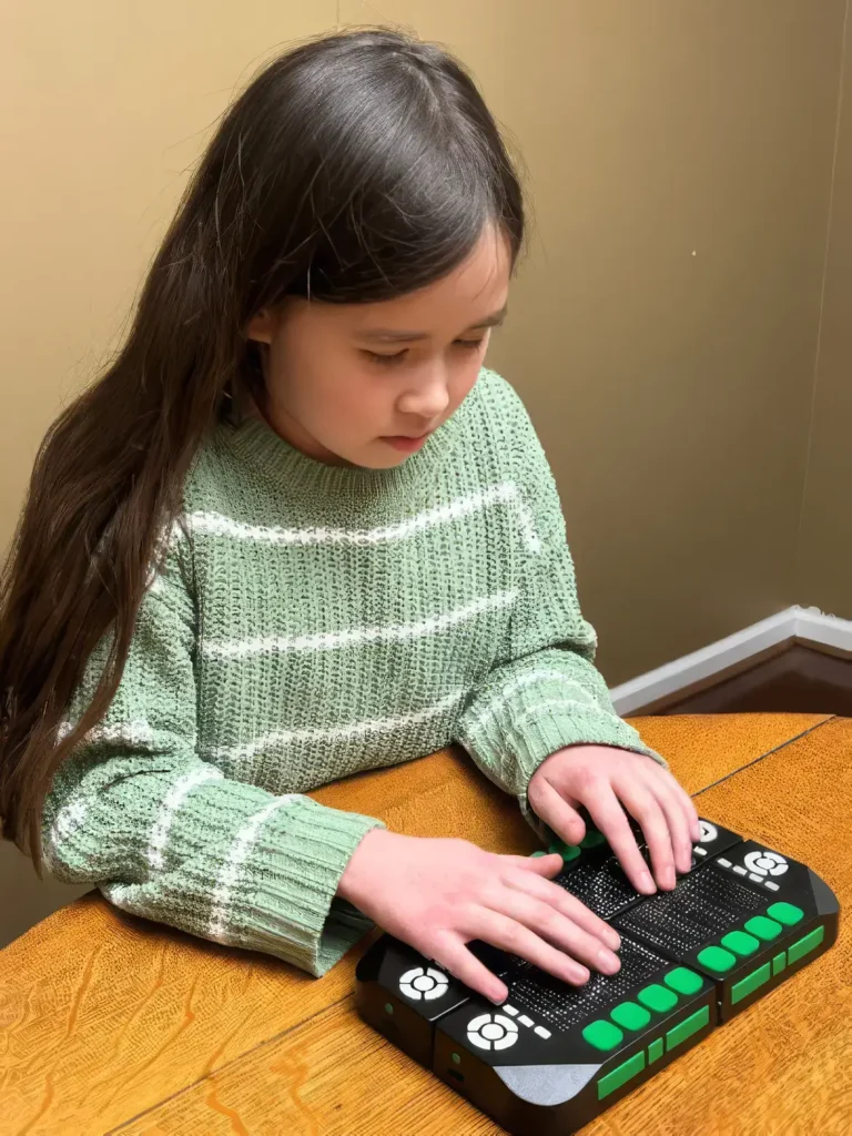 A young girl sitting at a wooden table, reading a tactile Braille display on a Cadence Workstation with her hands.