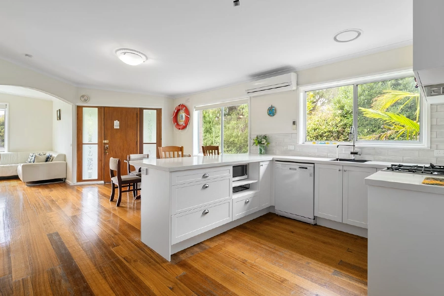 A bright open-plan kitchen and living area with white cabinetry, quartz countertops, a wooden dining table, timber flooring, and large windows overlooking greenery. A wooden front door with a decorative life-ring wreath is visible in the background, along with a cream sofa in the adjoining living room.