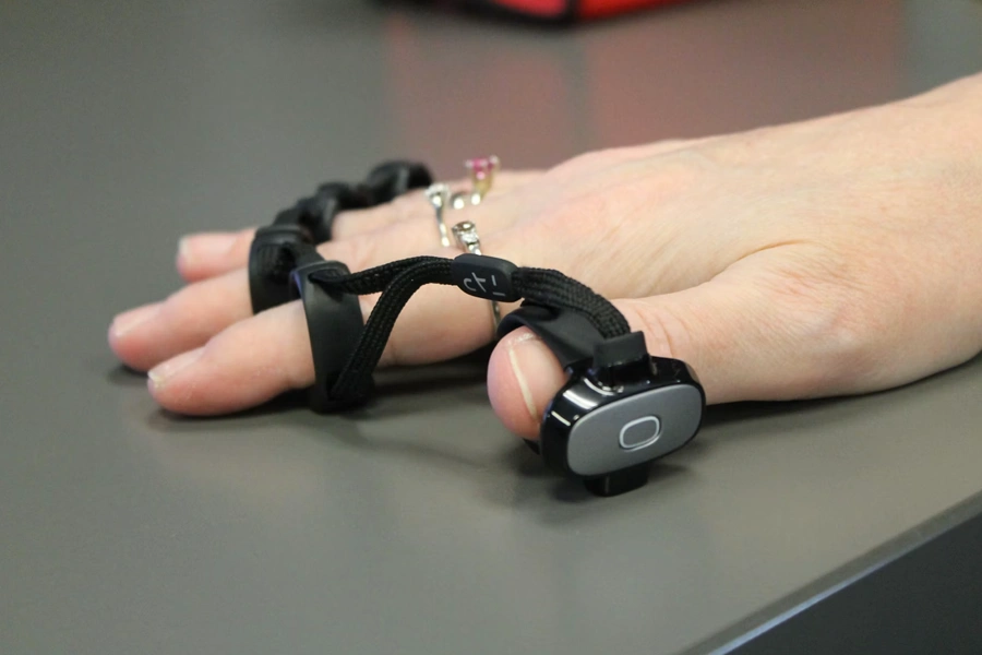 A close-up of a hand resting on a table wearing a wearable device with finger straps connecting to a small oval controller with a button, worn on the thumb, resting flat on a gray surface.