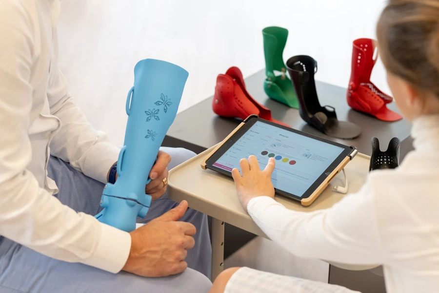 A clinician holds a light blue pediatric ankle-foot orthosis decorated with a butterfly pattern while a young patient touches a tablet displaying color and material customization options. Several other colorful orthotic leg braces — including green, black, and red designs — are arranged on a table in the background.