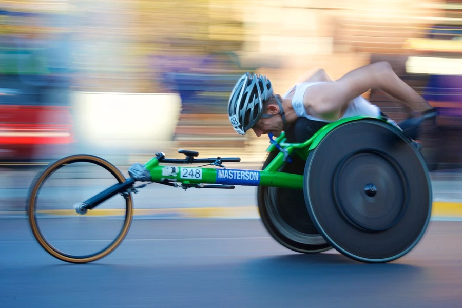 A wheelchair racer wearing a helmet and white tank top leans forward in an aerodynamic racing wheelchair, number 248 with "Masterson" printed on the frame, speeding down a road. The background is blurred with horizontal motion streaks, emphasizing speed, while the athlete and racing chair remain in sharp focus.
