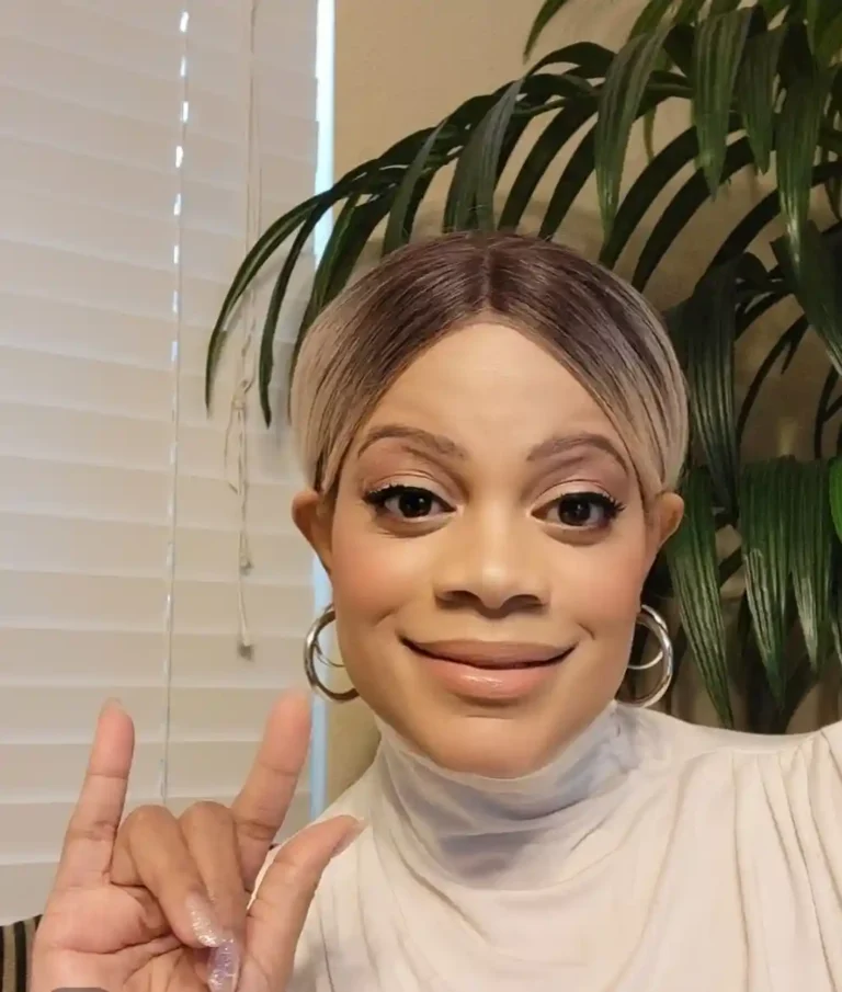 A smiling woman with blonde short hair and silver hoop earrings making the sign for "I love you" in American Sign Language.