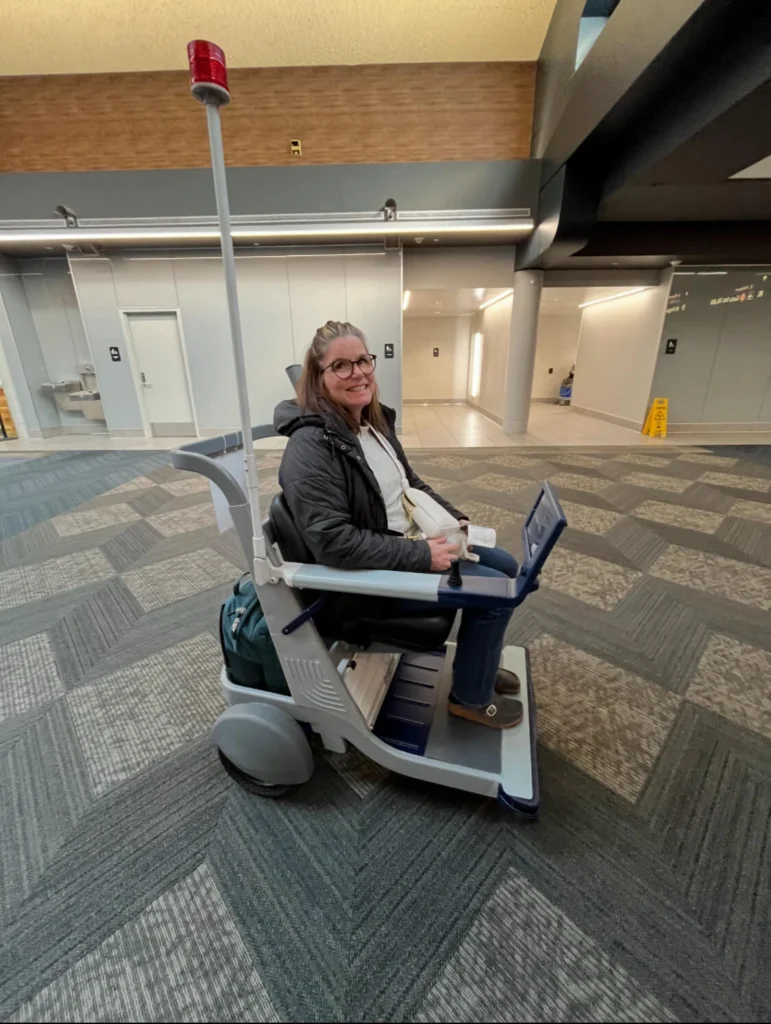 Three-quarter view of a smiling woman sitting in an autonomous mobility cart equipped with a high safety light pole inside an airport.