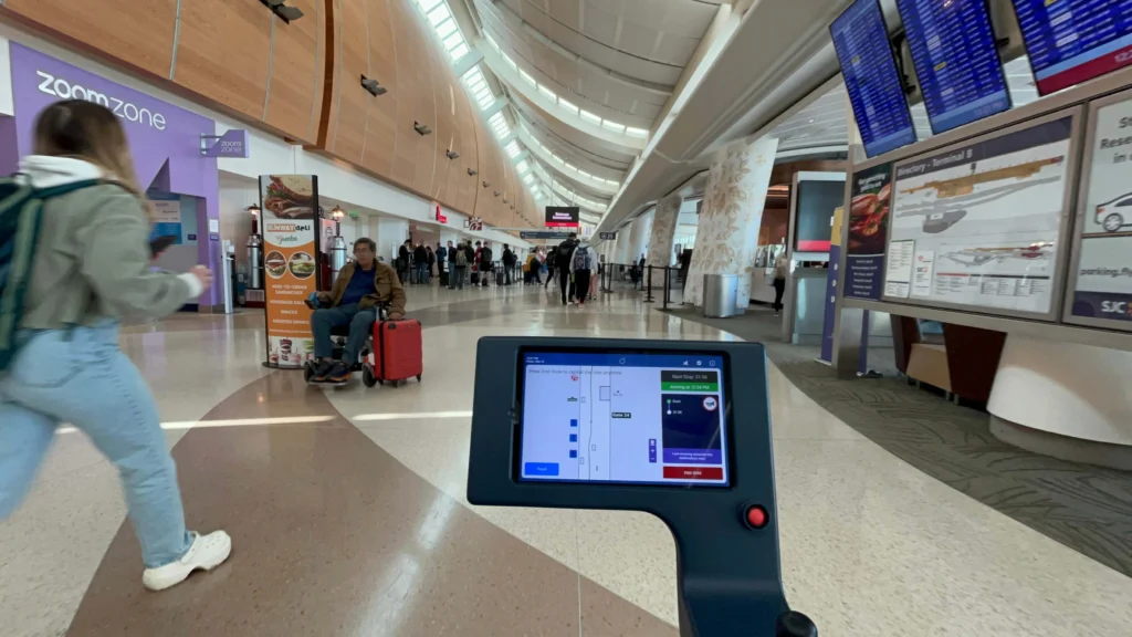 First-person view of the control screen on an autonomous mobility cart driving through a busy airport concourse.