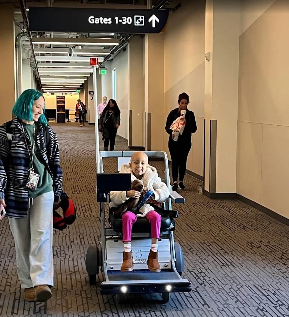 Front view of a child smiling while riding in an autonomous mobility cart down an airport hallway, accompanied by a person with teal hair walking alongside.