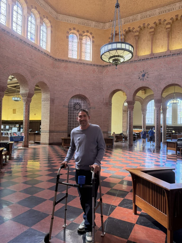 A man standing indoors holding onto a two-wheeled mobility walker on a red and black checkered tile floor inside a large, ornate hall with high arched doorways and a chandelier overhead.