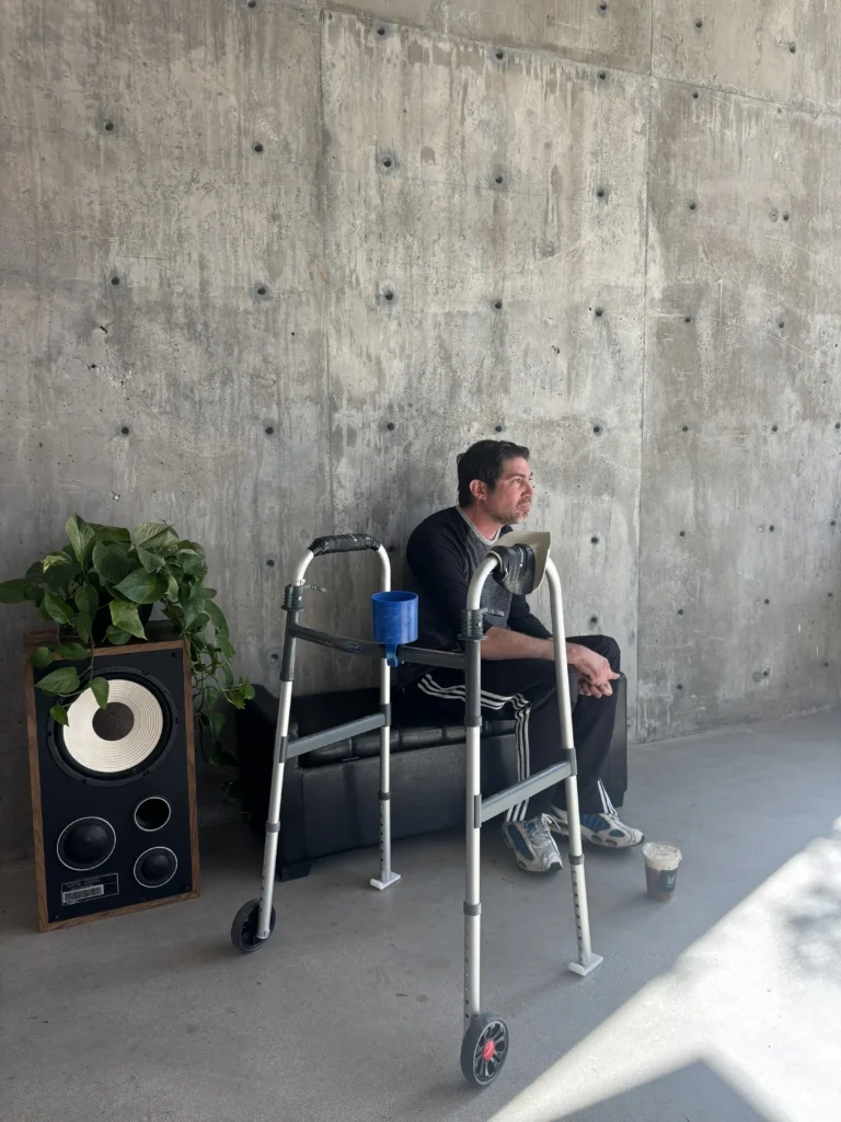 A man seated on a bench next to a mobility walker against a textured concrete wall, with a potted plant sitting atop a speaker nearby.