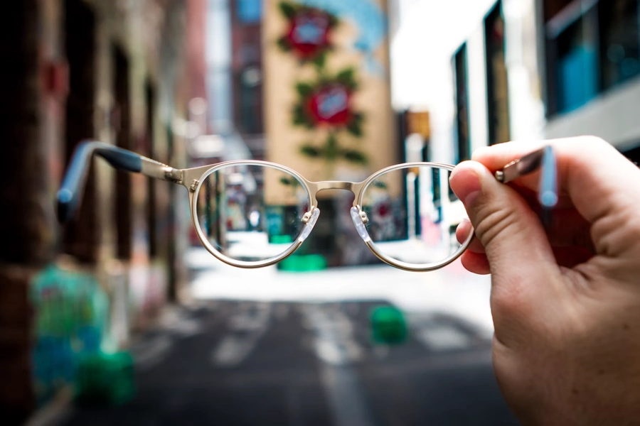 A hand holds up a pair of round wire-frame glasses toward the camera, with an urban alleyway featuring colorful graffiti and murals blurred sharply in the background, while the view through the lenses appears in focus.