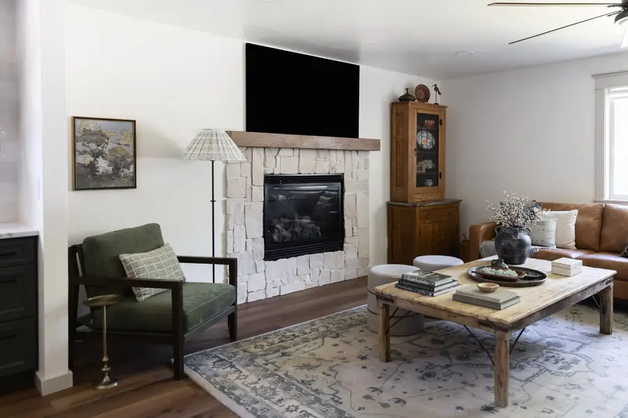 A cozy living room with a stone fireplace and mounted TV, a green armchair, wooden coffee table, leather sofa, and a vintage-style area rug.