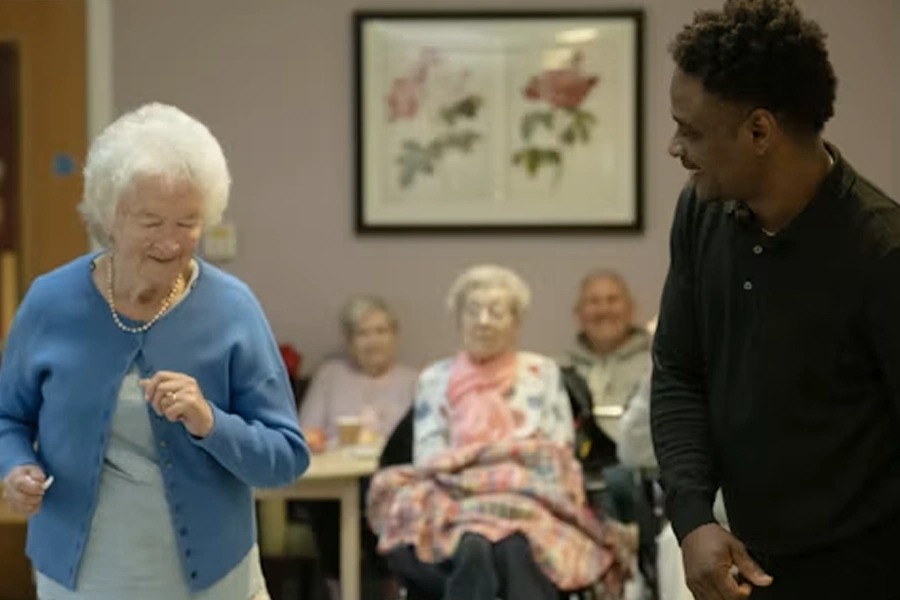 An elderly woman with white hair and a blue cardigan smiles and dances, while a younger man in a black shirt smiles and interacts with her, in what appears to be a care home common room with other elderly residents seated at a table in the background.