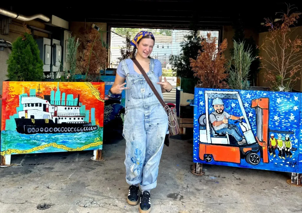 A woman in paint-splattered overalls making peace signs while standing between two large, hand-painted artwork planters outdoors.