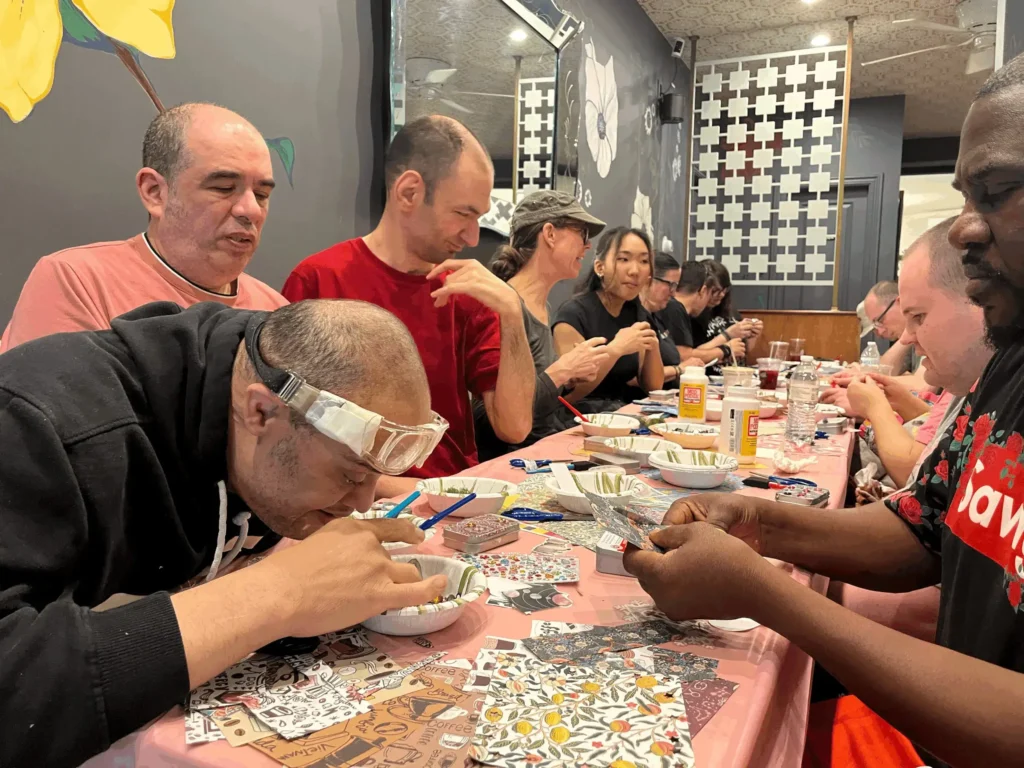 A group of people seated around a pink table working on craft projects with glue, patterned paper, and tools.