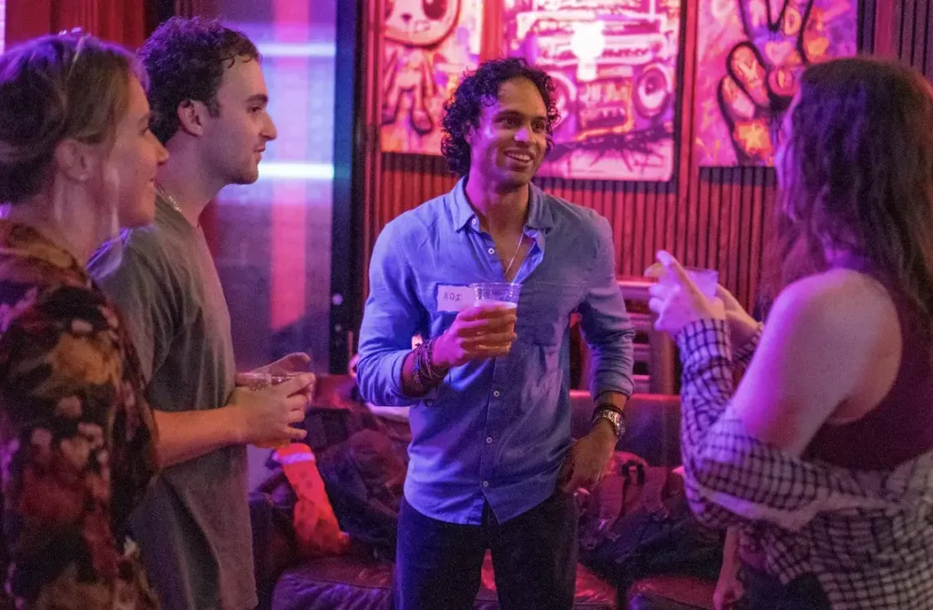 A smiling man wearing a blue shirt and name tag holding a drink and talking to people at a dimly lit, neon-lit gathering.