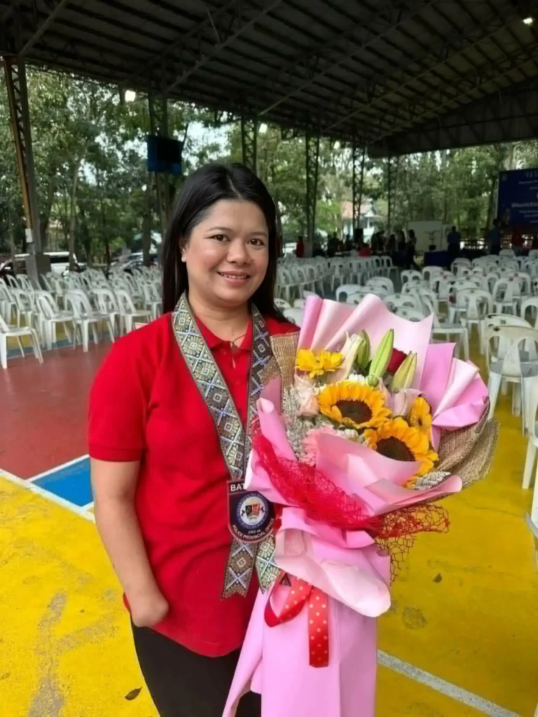 Chef Maricel Apatan in a red shirt wearing an honor sash and holding a large bouquet of pink paper-wrapped sunflowers and roses in an outdoor cour