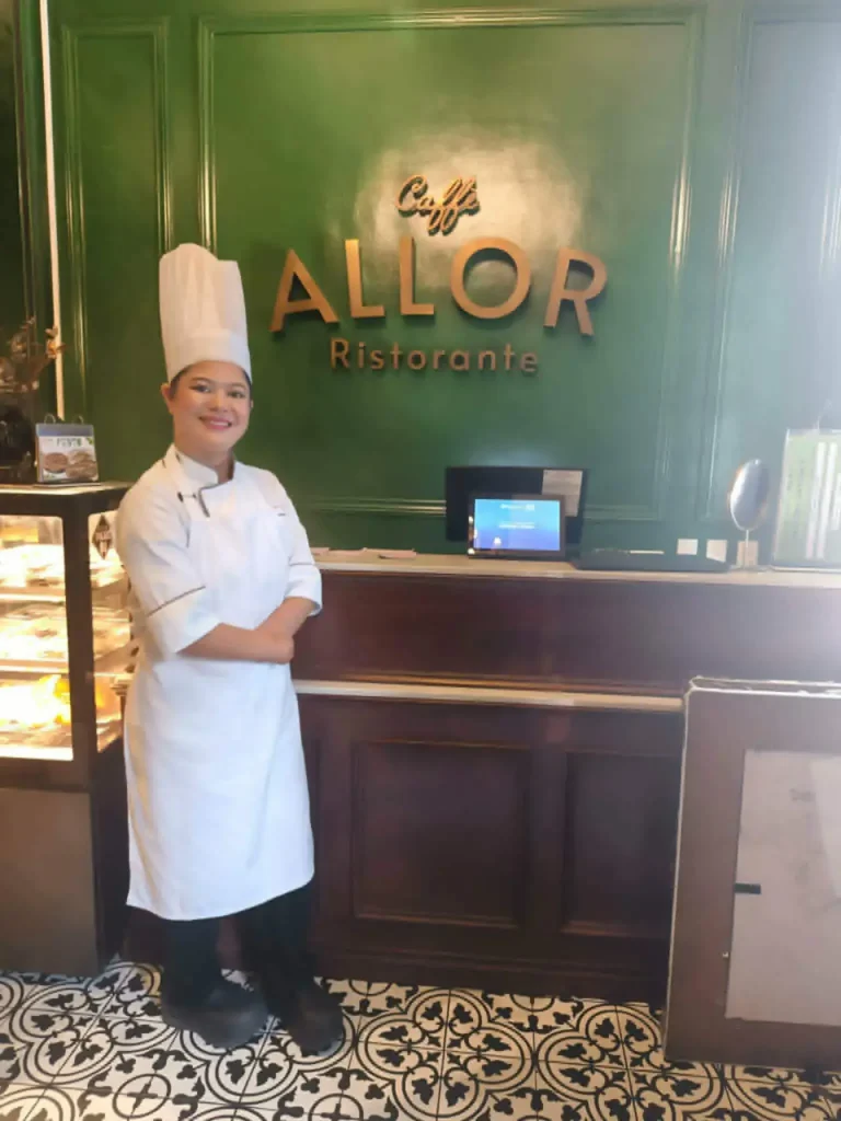 Chef Maricel Apatan in a white toque and chef coat posing by the wooden counter inside Caffè ALLOR Ristorante.