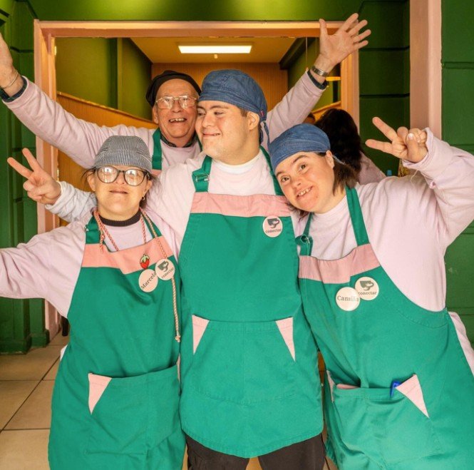 Four people wearing matching teal aprons and caps pose playfully together in a doorway, some making peace signs, in front of a bright green wall.