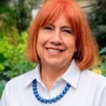 Outdoor portrait of a smiling woman with shoulder-length auburn hair, wearing a white collared shirt and a chunky blue beaded necklace, with a blurred green garden background