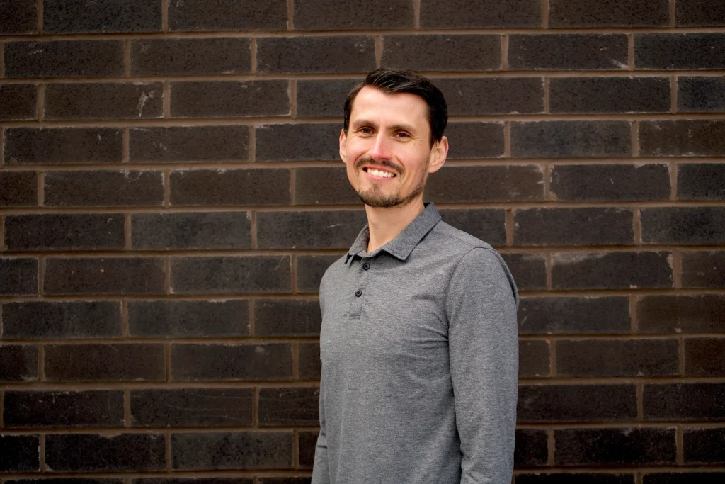 A smiling man with dark hair and a light beard standing in front of a dark brick wall, wearing a grey polo-style long-sleeve shirt.