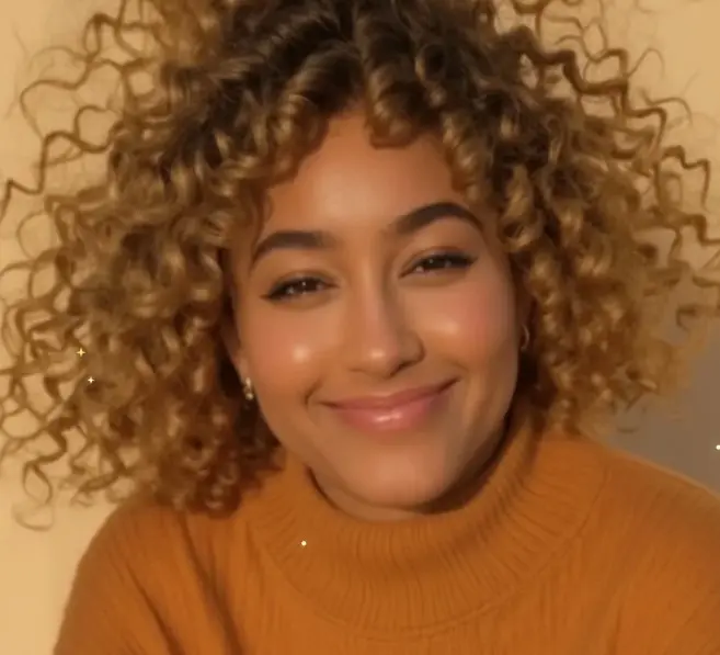Close-up portrait of a smiling young woman with voluminous blonde curly hair wearing a mustard-yellow turtleneck sweater.