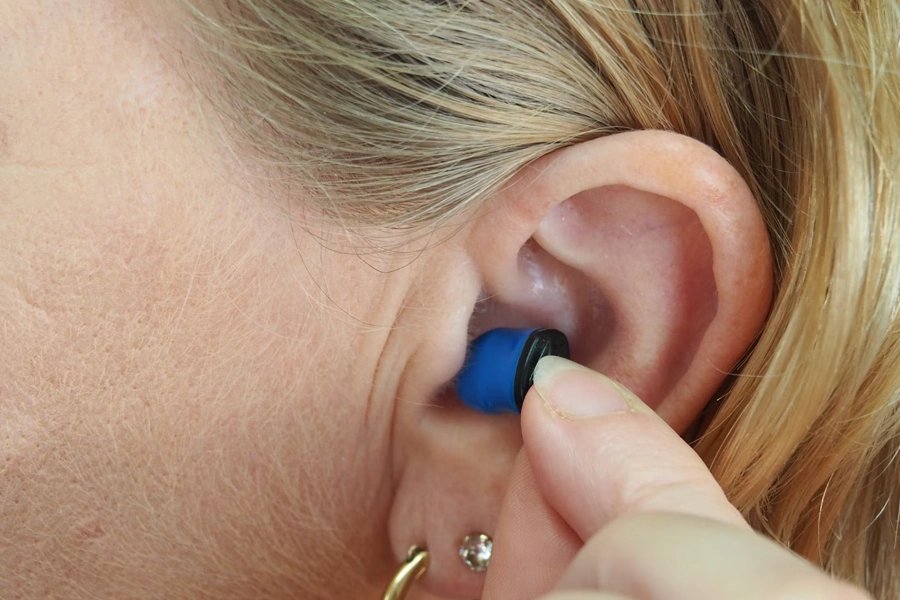 Close-up of a person with blonde hair inserting a blue and black earplug or in-ear device into their ear canal.
