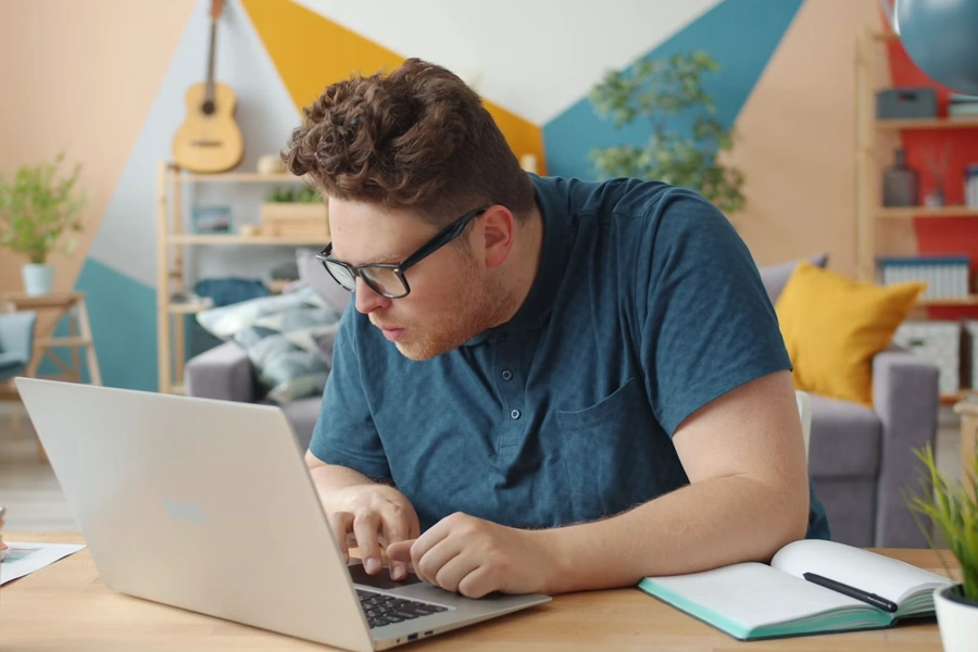 A young man with curly hair and glasses focuses intently while typing on a laptop at a wooden desk, with an open notebook and pen beside him, in front of a colorful geometric-patterned wall and a living room with a guitar, shelves, and a gray sofa in the background.