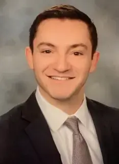 A headshot portrait of a smiling man with short brown hair wearing a dark suit jacket, white dress shirt, and light grey tie.