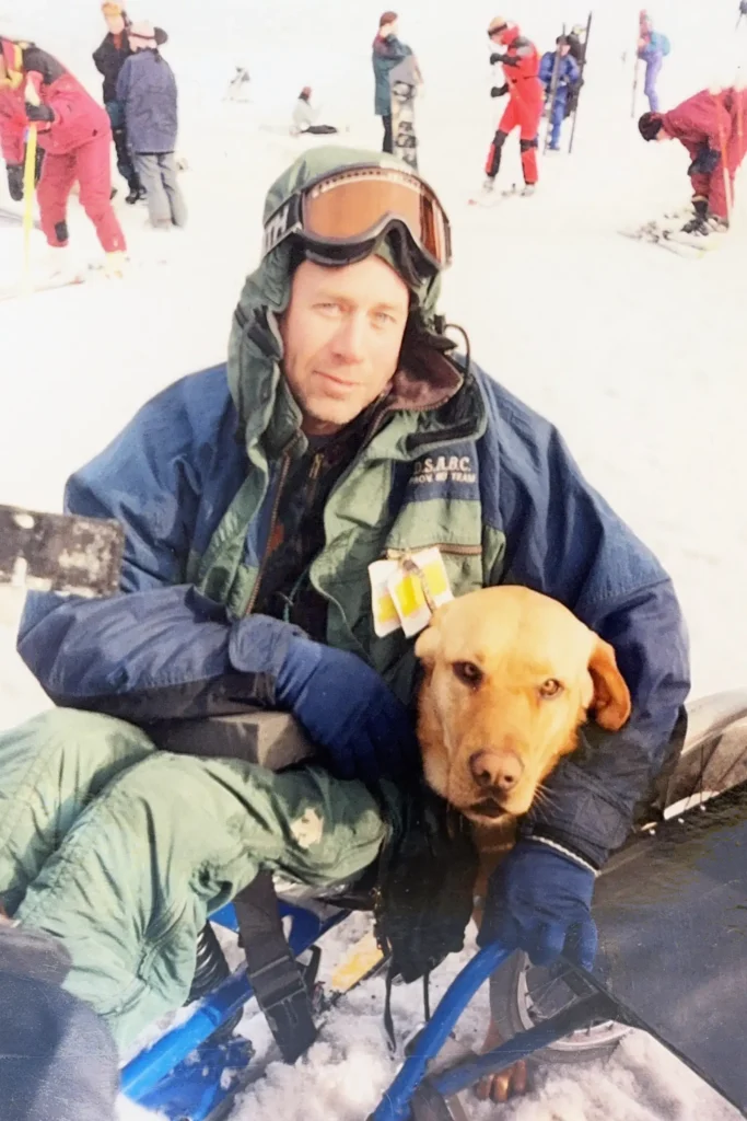 A close-up portrait of a sit-skier bundled up in ski gear resting on the snow while hugging a golden retriever close to his side.