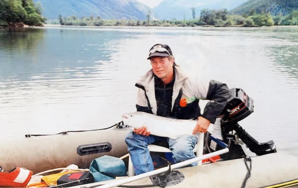 A man sitting in an inflatable motorized boat holds a large silver fish on his lap while floating on a calm river surrounded by mountains and trees.