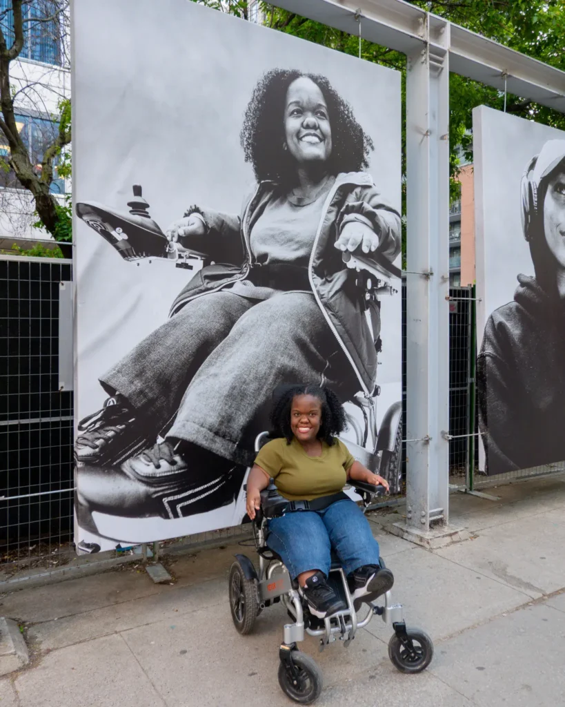 A smiling Black woman in a olive green shirt sits in her motorized wheelchair outdoors in front of a large, black-and-white billboard featuring a photograph of herself.