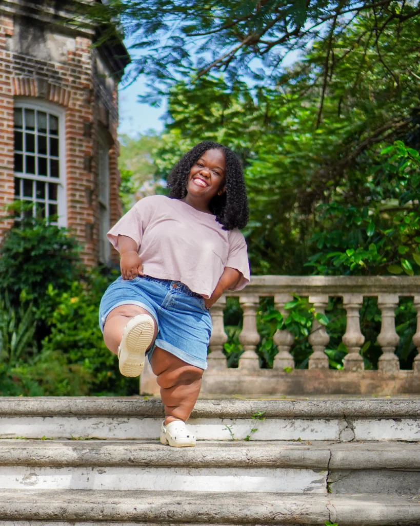 A smiling Black woman with curly hair poses playfully on outdoor stone steps, lifting one leg in a crop top, denim shorts, and clogs.