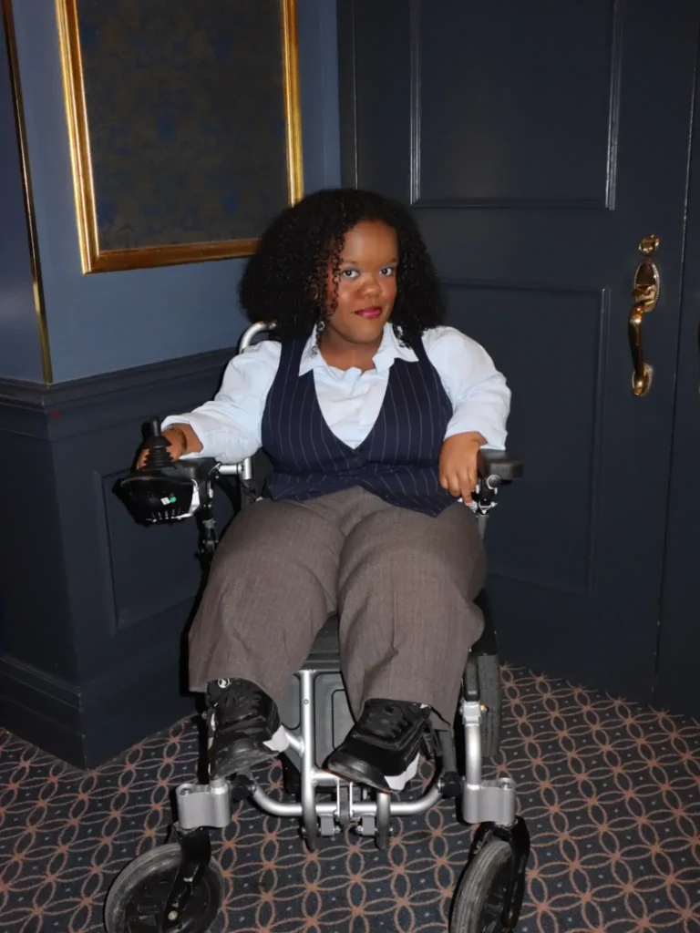 A Black woman sitting in a motorized wheelchair indoors, wearing a light blue button-up shirt, a pinstriped vest, gray trousers, and dark sneakers.