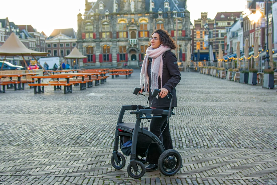 A woman wearing a scarf and coat walks with a modern rollator (wheeled walker) across a cobblestone town square, with historic European buildings and a market stall in the background at dusk.