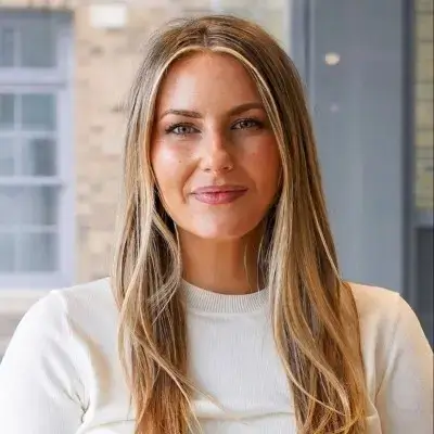 A close-up headshot of a smiling woman with long light-brown hair, subtle blonde highlights, and a white top.