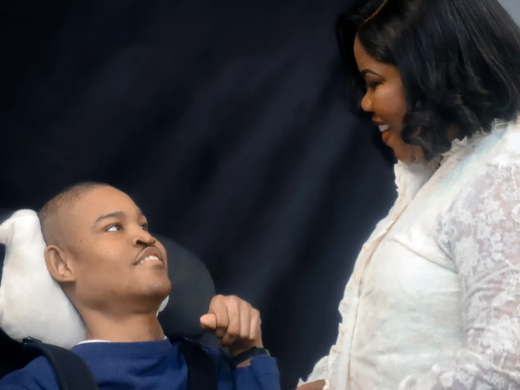 A woman in a white lace top looks fondly and smiles down at a young man resting his head in a wheelchair against a dark backdrop.