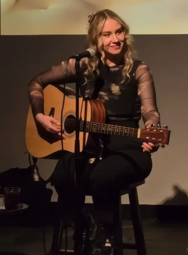 A smiling woman with blonde hair sitting on a stool onstage and playing an acoustic guitar.