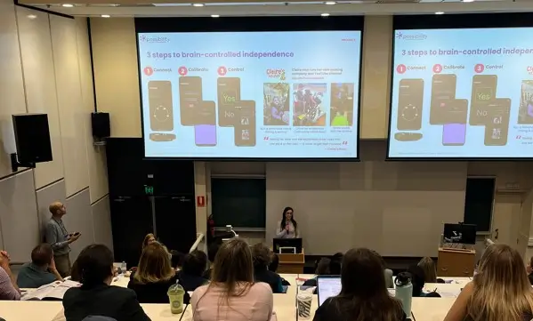 A speaker standing on stage in a lecture hall giving a presentation on brain-controlled independence with slides projected on large screens.