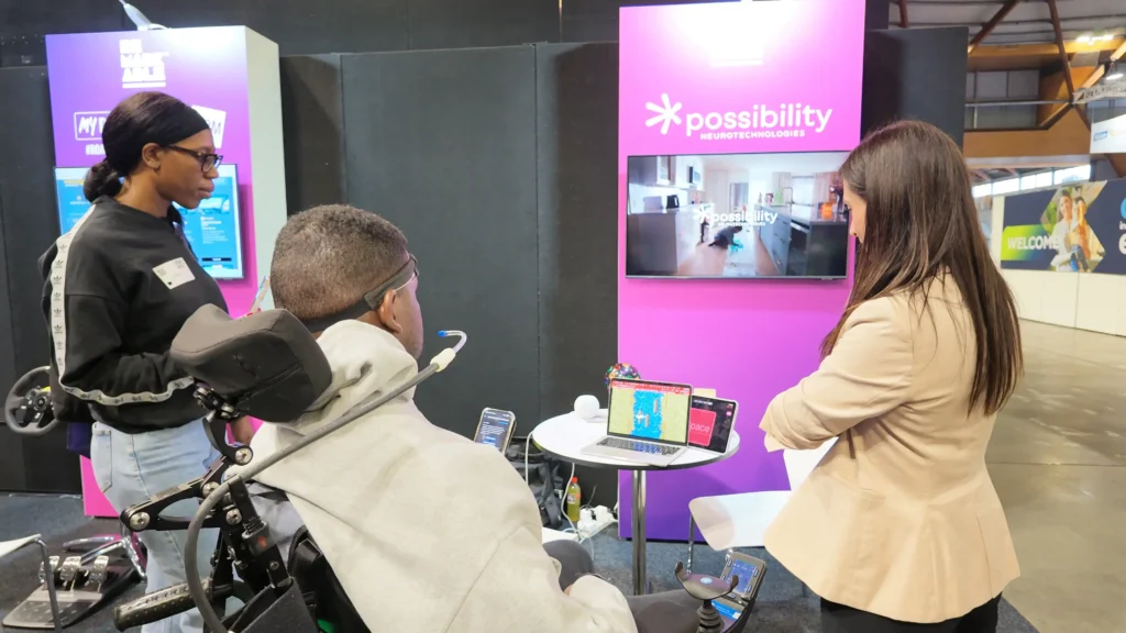 A woman presenting a laptop and tablet demo at a Possibility Neurotechnologies exhibition booth to a person sitting in a wheelchair.