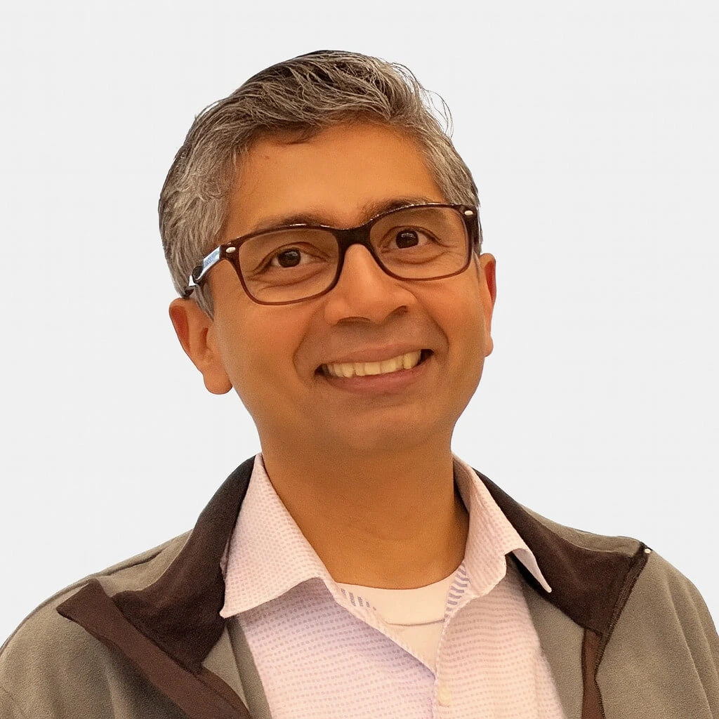 Headshot of a smiling man with glasses, short salt-and-pepper hair, wearing a white collared shirt under a gray jacket.
