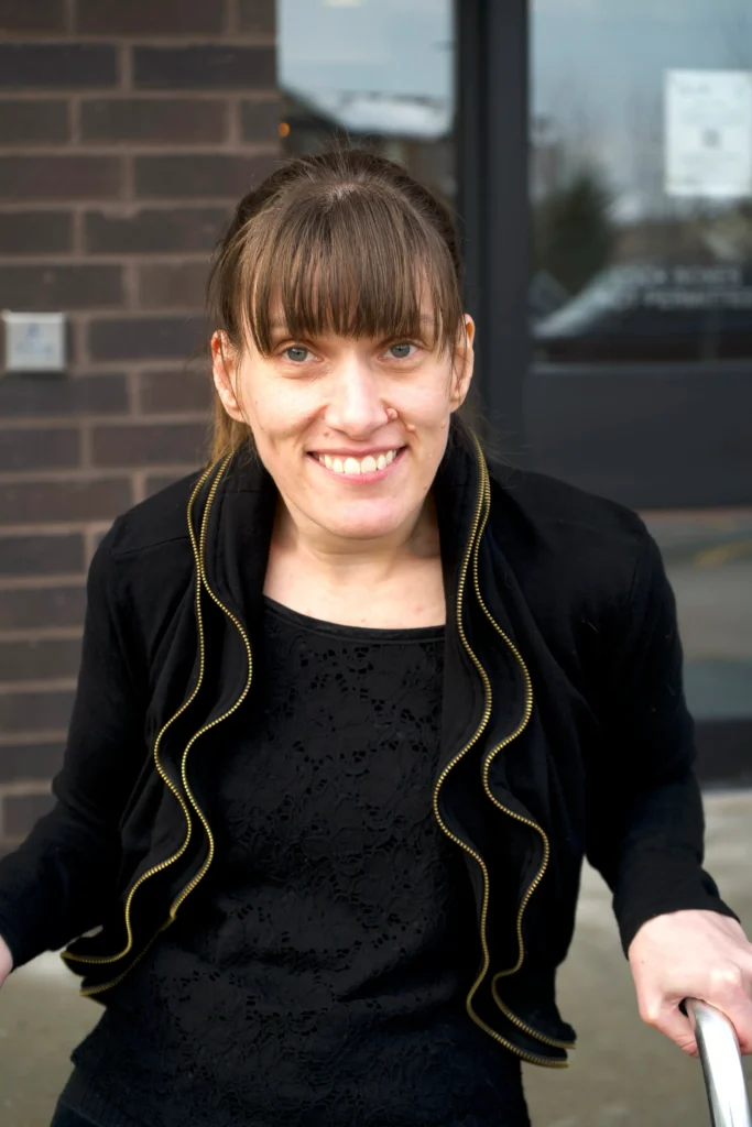 A smiling woman with brown hair and bangs leaning forward outside a building entrance with dark brick and glass doors, wearing a black jacket with wavy trim over a patterned black top.