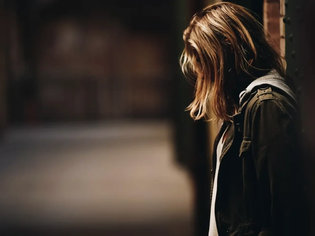 A woman with long blonde-highlighted hair stands with her head bowed, face obscured by her hair, leaning against a dark doorway at the end of a dim, shadowy corridor.