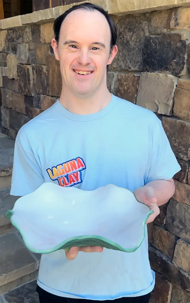 A smiling man holding up a finished light blue ceramic bowl with a wavy green rim.