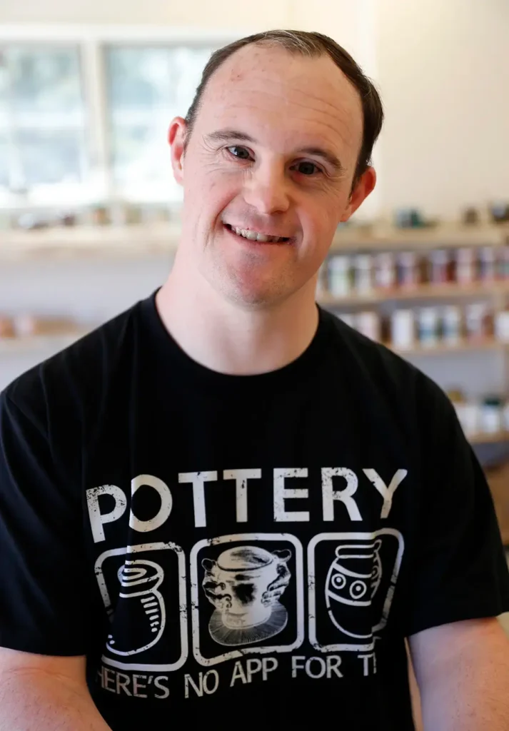 A close-up portrait of a smiling man wearing a black t-shirt with pottery graphics.