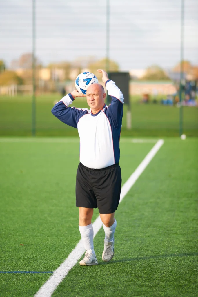 An older man preparing to make a throw-in on a green soccer pitch while wearing sportswear.