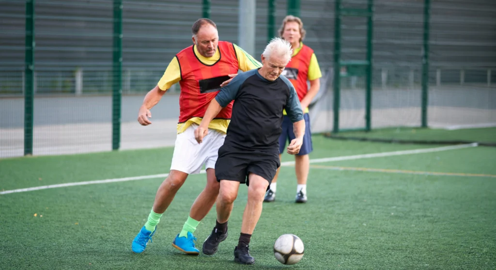 Senior men playing soccer on an outdoor turf field, with one controlling the ball near the opponent.