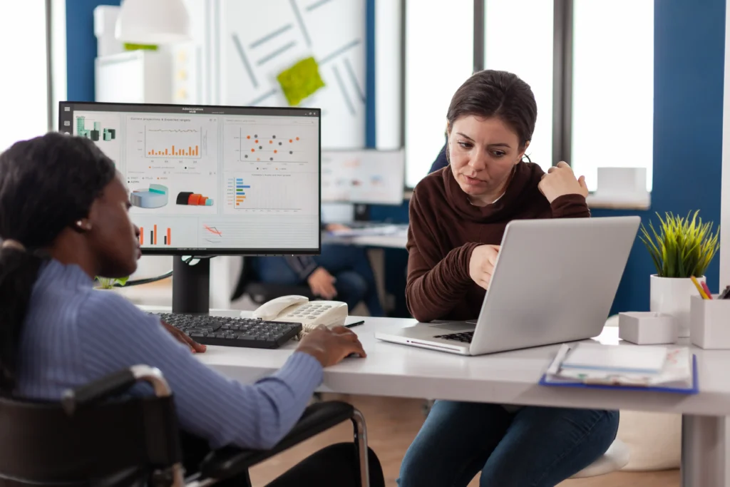 Two women collaborating at a office desk in front of a laptop and desktop computer displaying data charts.
