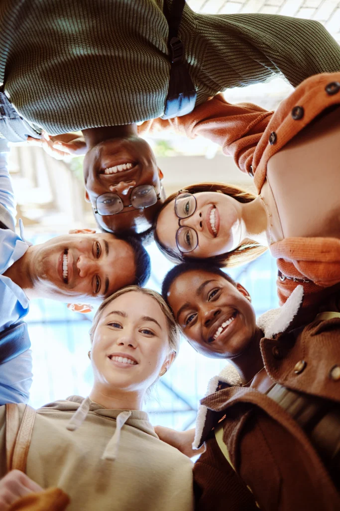 Low-angle view of five diverse students standing in a circle, smiling down at the camera with their heads together.