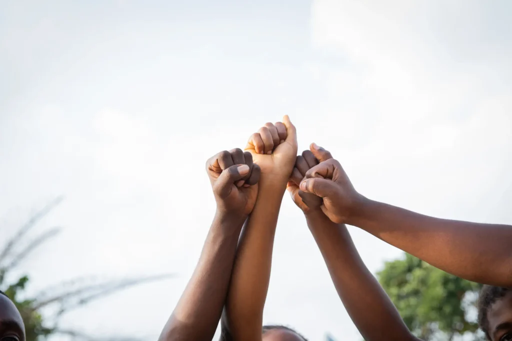 A low-angle shot of several hands raised in the air with clenched fists in a show of unity and solidarity against a bright sky background.